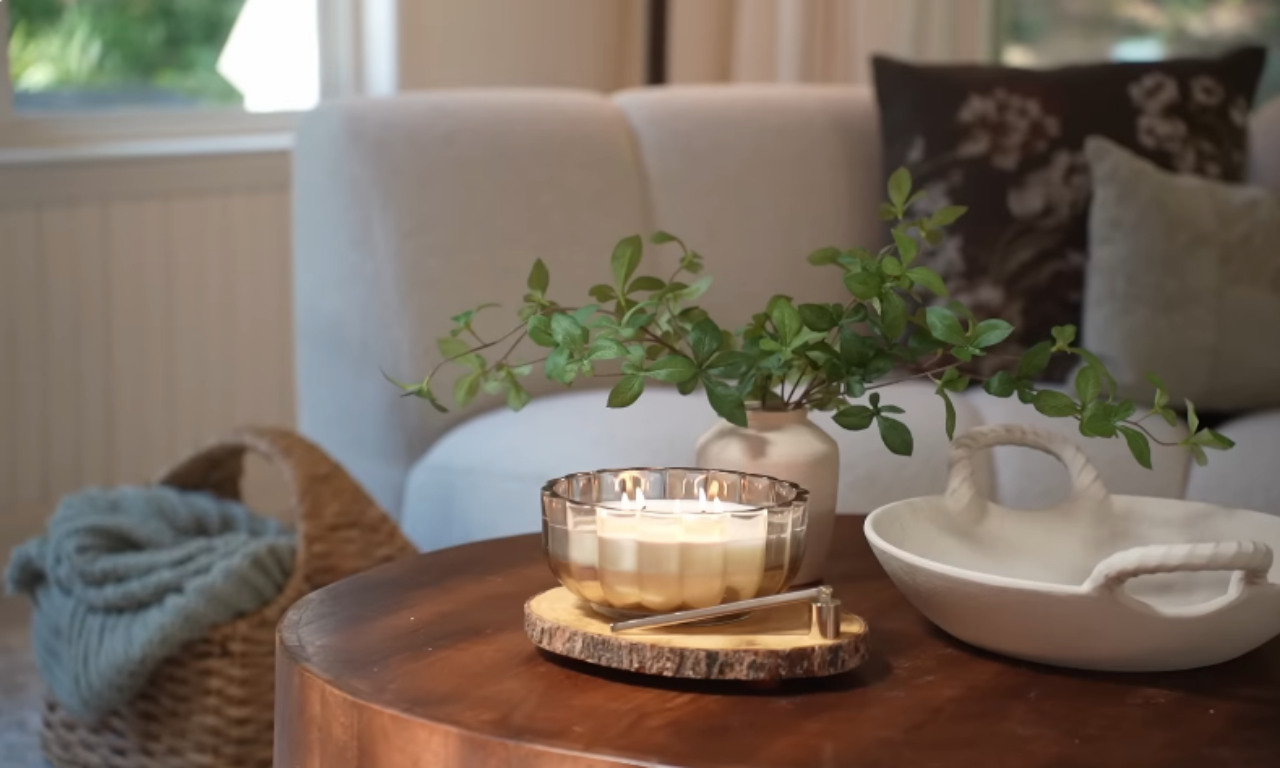 A cozy coffee table setting featuring a lit multi-wick candle in a glass container, a white ceramic tray, a vase with green leafy branches, and a candle snuffer on a wooden coaster. The background includes a white couch with a dark floral cushion and a woven basket holding a blue blanket.