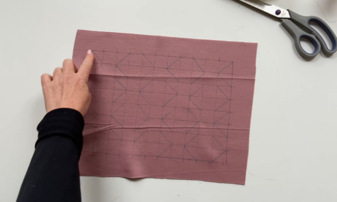 A hand points to a piece of dusty rose-colored fabric that has been marked with a grid and diagonal lines forming a geometric pattern. The fabric is laid flat on a white surface next to a pair of scissors.