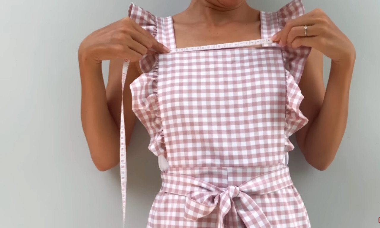 Woman wearing a pink gingham dress with ruffled shoulder straps, using a measuring tape to measure the width of the bodice across the chest.
