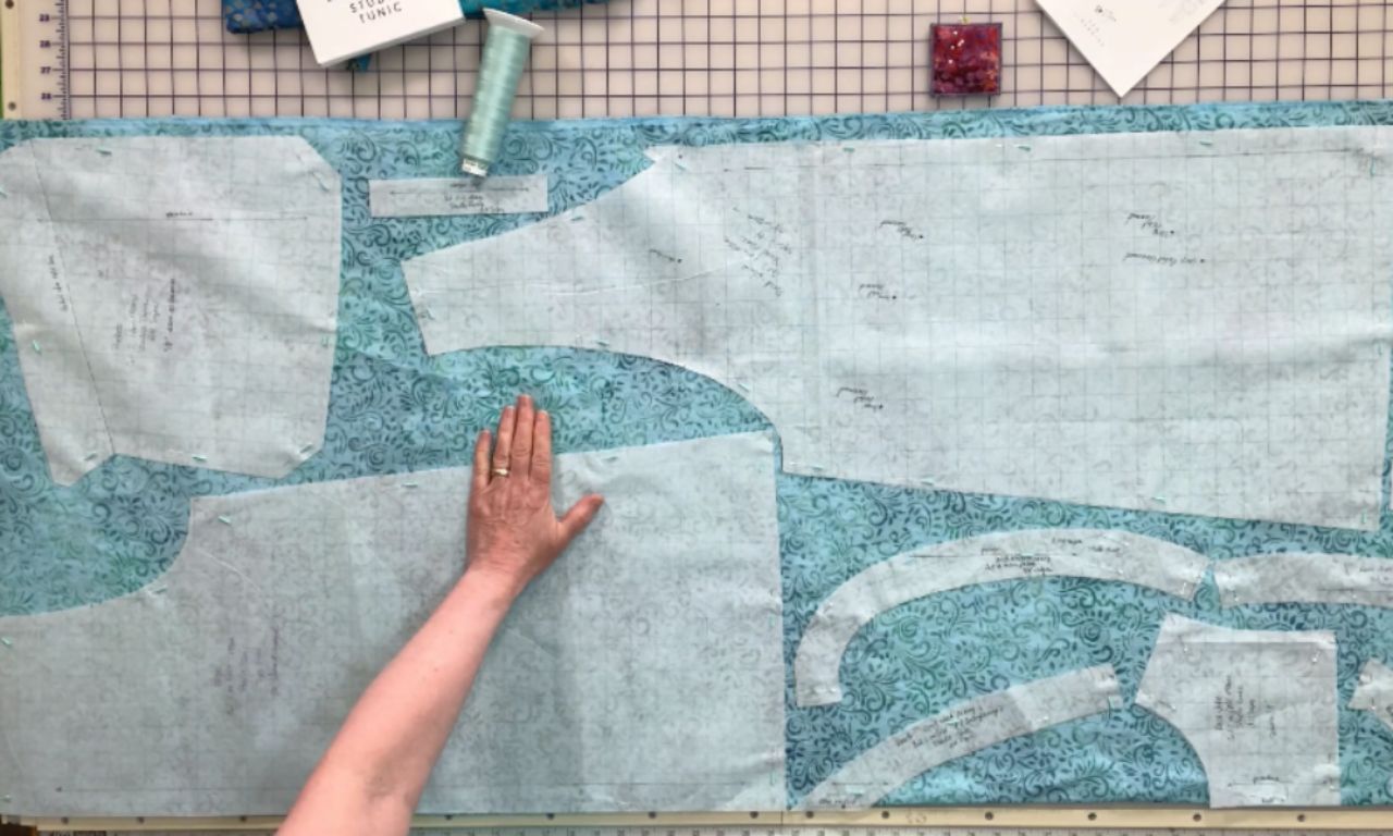 A hand lays flat on blue patterned fabric with multiple garment pattern pieces pinned to it, ready for cutting. The background includes a gridded cutting mat, sewing supplies, and thread spools.