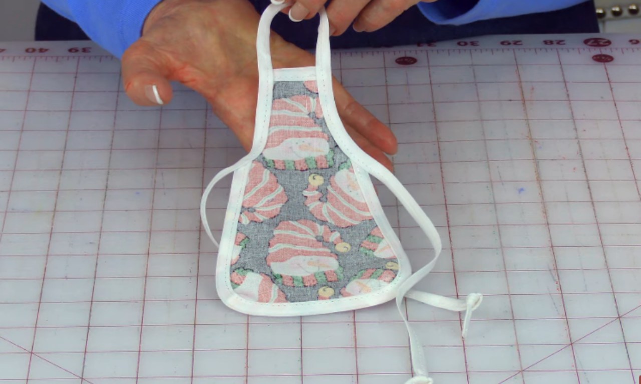 Close-up of a person holding a small, finished fabric apron with white trim and a pink-and-gray cupcake print. The apron appears to be designed for a wine or olive oil bottle.
