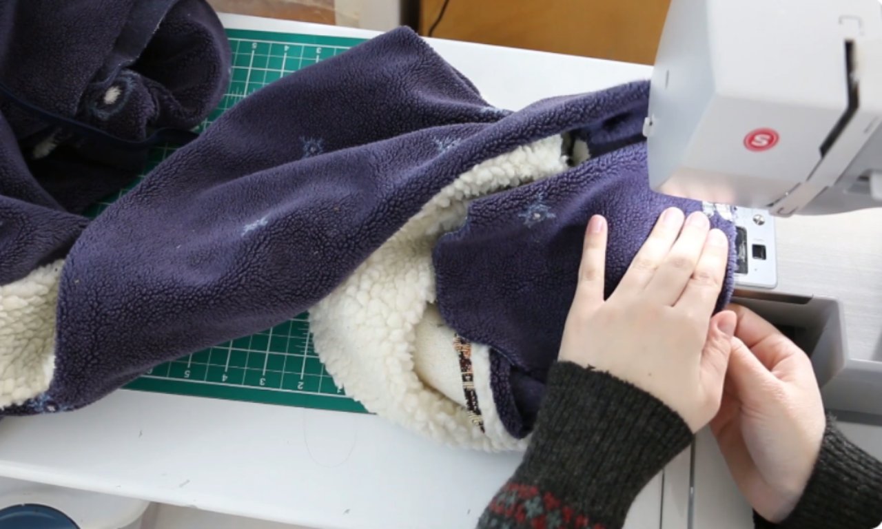 Close-up of hands sewing a navy blue fleece fabric with a sherpa lining using a sewing machine. The fabric features small white snowflake-like motifs and is partially turned inside out.