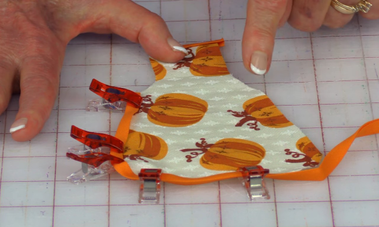 A small fabric apron featuring an orange pumpkin pattern is laid flat on a cutting mat. The apron is partially constructed and secured with red sewing clips, with an orange trim being attached.