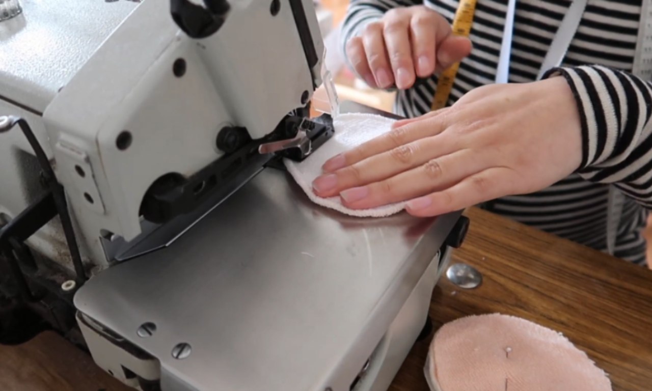 A person using a sewing machine to stitch the edges of a round, reusable fabric pad. The person is wearing a striped shirt and a yellow measuring tape is draped around their neck. Partially sewn pads are visible on the table.