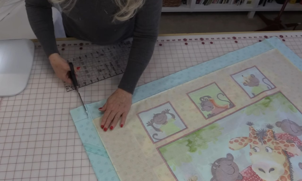 A person is using scissors to trim the edges of a quilt that is laid out face-down on a cutting mat. The quilt has a light blue and beige border and features illustrations of cartoon monkeys, a giraffe, and other animals. A clear quilting ruler is placed under the scissors for guidance.