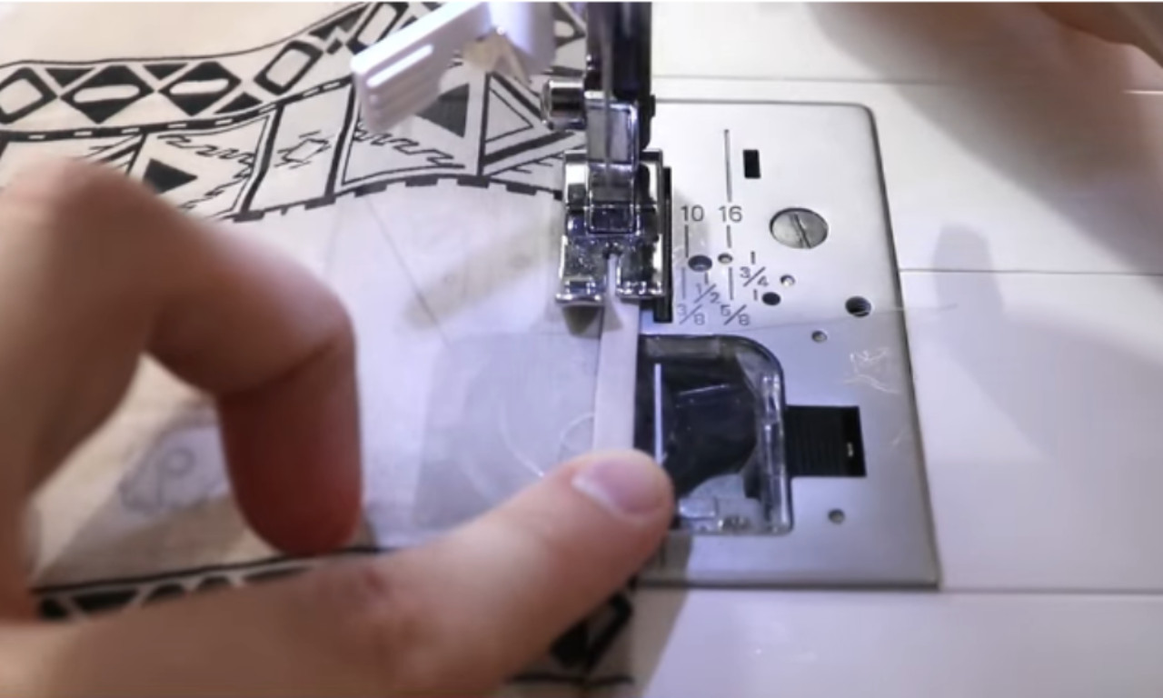 A close-up view of a sewing machine stitching the edge of a patterned fabric. A hand is guiding the fabric under the presser foot as the machine sews along the seam. The fabric has a black and white geometric design.