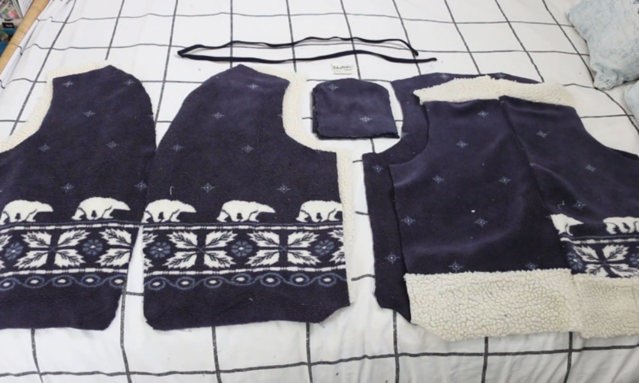 Multiple cut fabric pieces laid out on a bed with a grid-patterned blanket, showing the components for sewing a sherpa-lined vest. The pieces include navy fleece with polar bear and snowflake motifs, along with a drawstring.