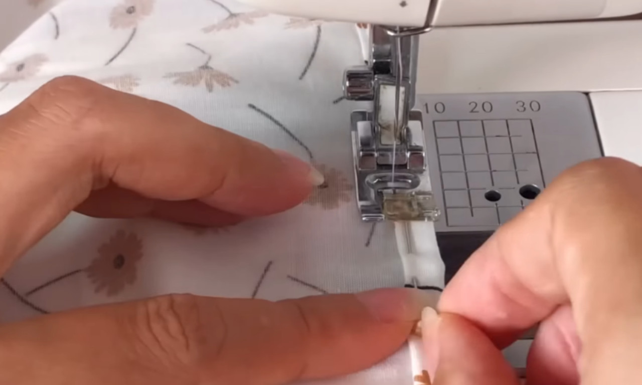 Close-up view of hands guiding fabric with a floral print through a sewing machine, stitching a narrow fold along the edge of the material.