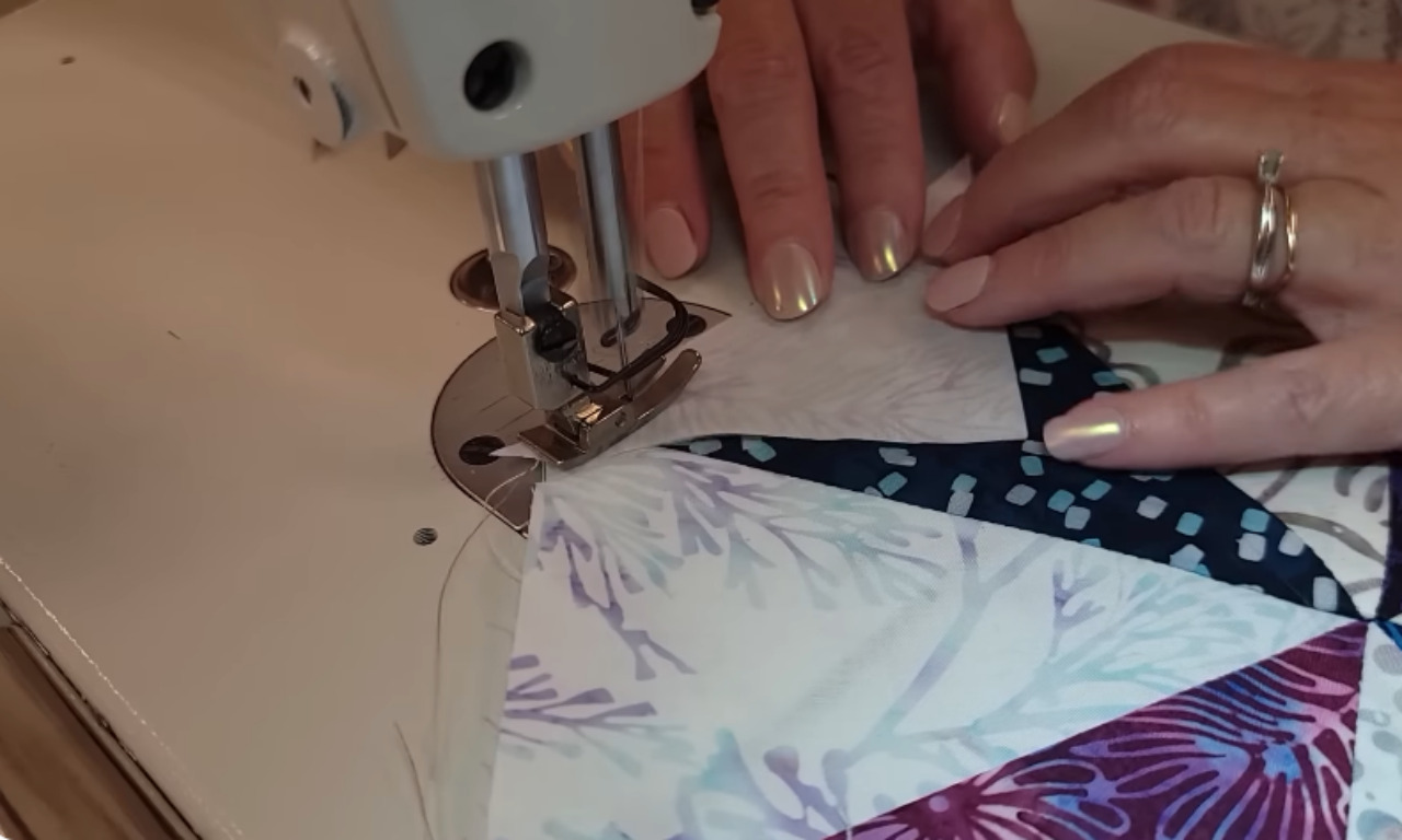 A close-up view of hands guiding colorful triangle-shaped fabric pieces through a sewing machine, showing the precision of stitching in the quilt-making process.