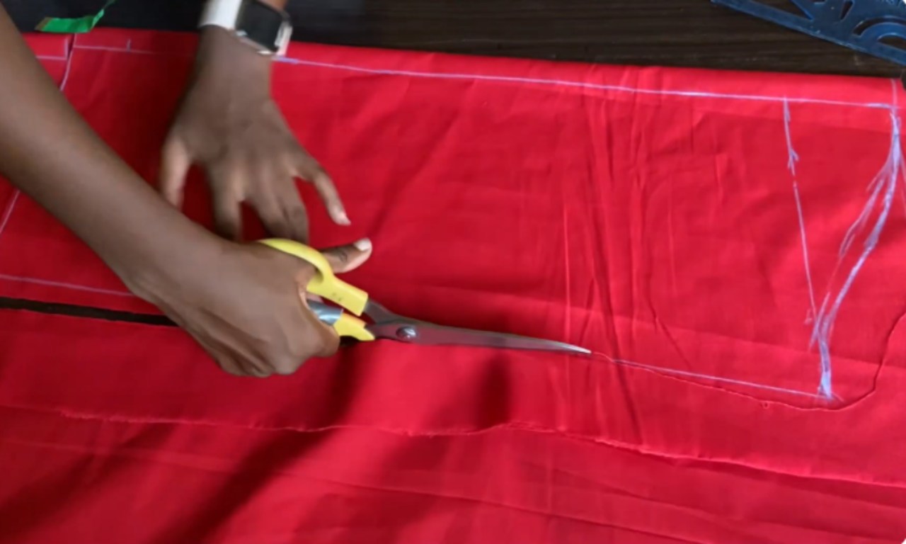 A person using large yellow-handled fabric scissors to cut along white chalk lines on bright red fabric. The lines outline the pattern for a split sleeve piece, indicating the initial cutting stage in the sewing process.