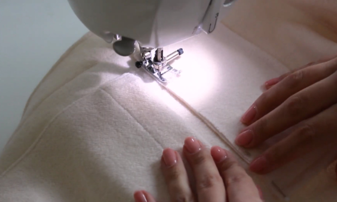 Close-up of a sewing machine stitching a seam on light beige fabric. A person's hands are gently guiding the material under the needle, indicating careful sewing work in progress.