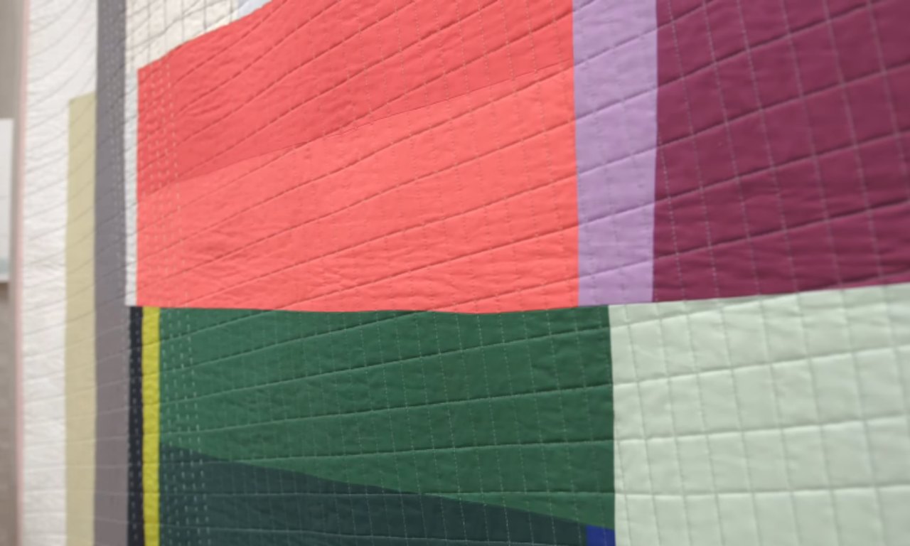 A modern quilt with large, solid color blocks in pink, burgundy, lavender, green, and cream, arranged in geometric shapes with diagonal and vertical quilting lines.
