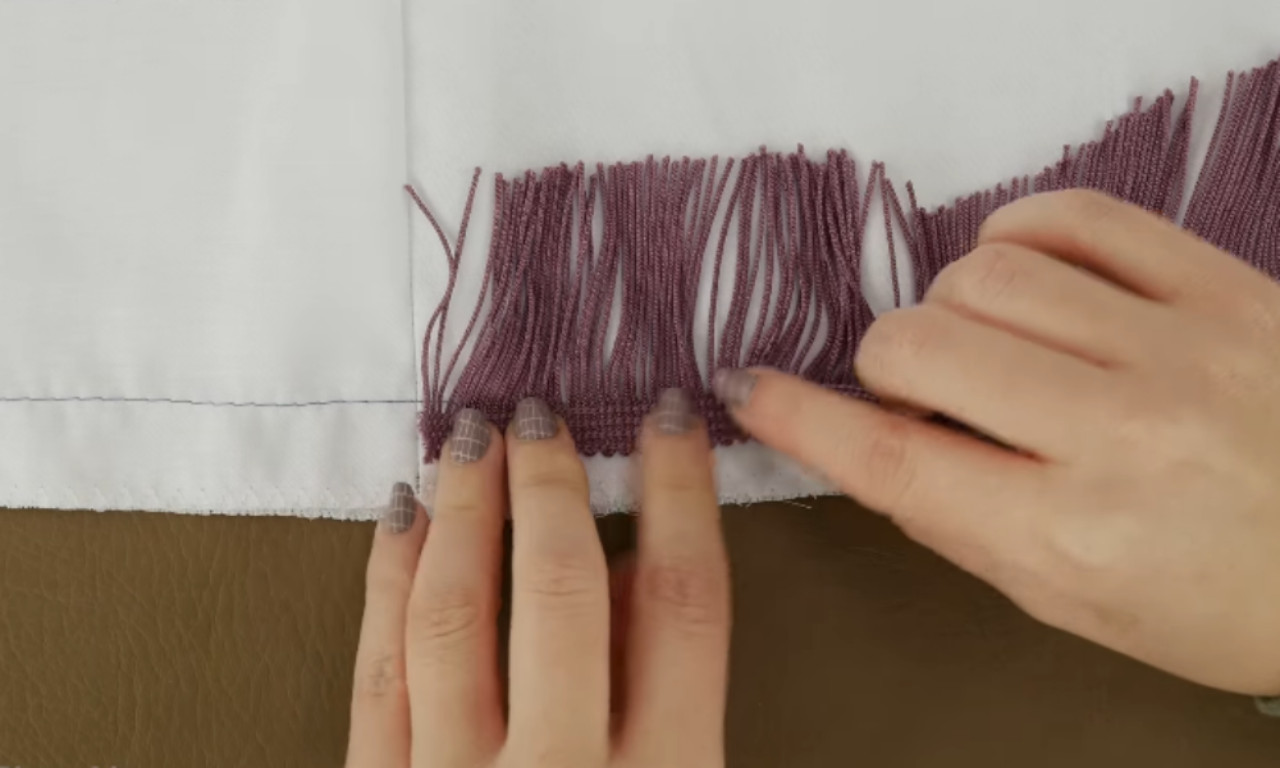Hands smoothing and adjusting mauve fringe that has been sewn onto the edge of white fabric, ensuring it lays flat and even.