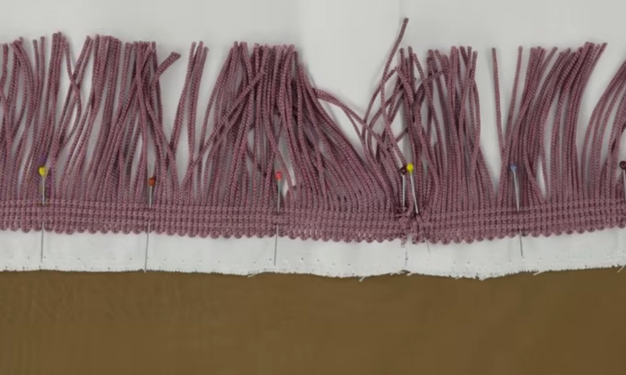 Close-up of mauve fringe trim pinned to the edge of a piece of white fabric using colorful sewing pins, ready for stitching.