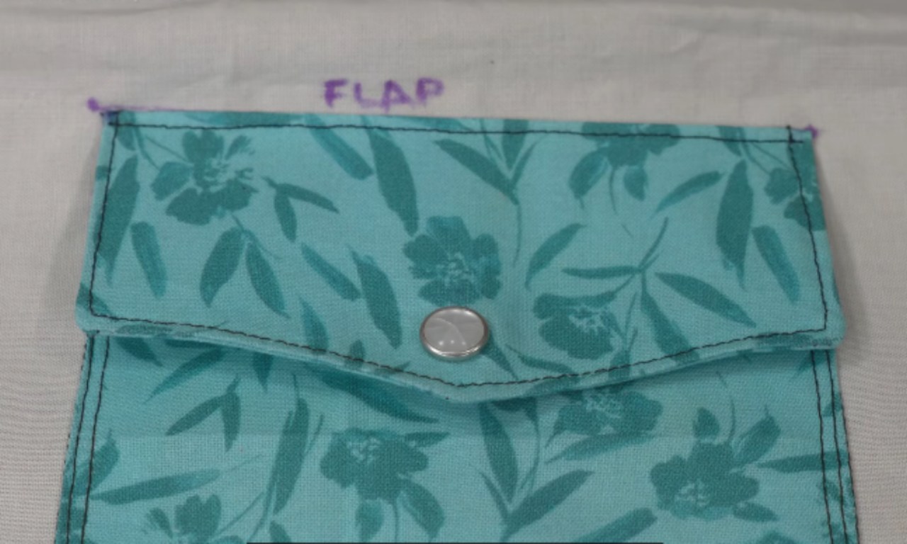 A close-up view of a fabric pocket with a flap sewn onto a light-colored background. The pocket is made from teal fabric with a floral pattern, and features black topstitching and a metal snap fastener in the center of the flap. The word "FLAP" is marked above the pocket in purple ink.