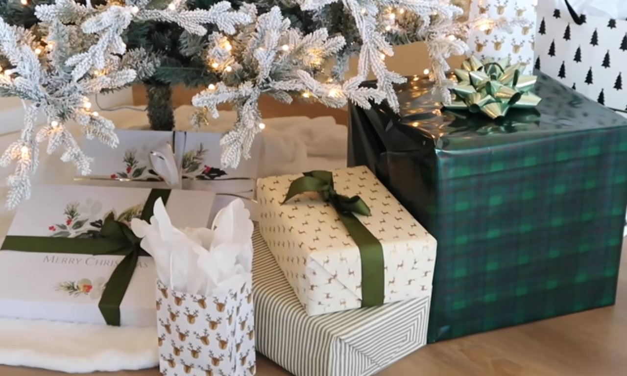An assortment of wrapped Christmas gifts sits beneath a flocked Christmas tree. The presents are wrapped in various papers, including a green and blue plaid, a white paper with deer and trees, and a white paper with green bows and branches.