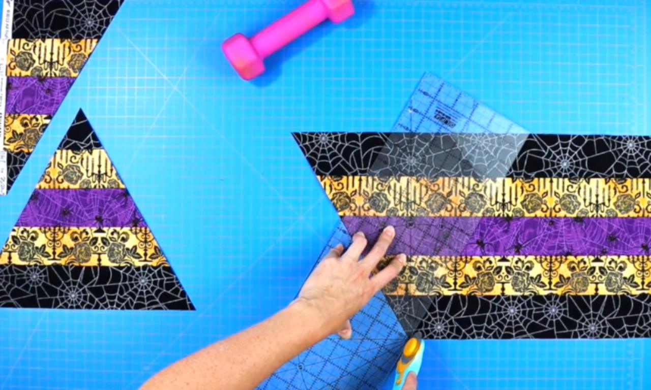 A completed hexagon table topper made with Halloween-themed fabric strips in black, gold, and purple. The fabric has patterns like spider webs, skulls, and roses. The topper is placed on a gray felt pad over a blue cutting mat. Quilting tools such as a ruler, rotary cutter, scissors, and fabric clips are visible on the side.