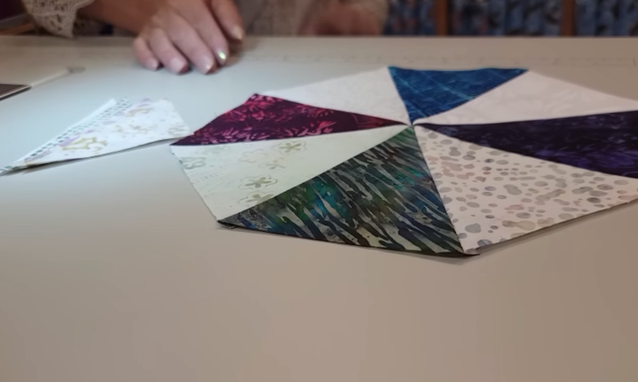 A partially assembled quilt block with multiple triangle pieces arranged into a circular star pattern is laid out on a table, with a person’s hands nearby, preparing the next steps in the layout.