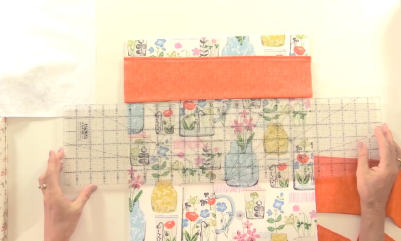 A person measuring and aligning colorful floral fabric with a transparent quilting ruler on a white surface. Orange and white fabric pieces are folded and arranged above the floral fabric. The scene is well-lit and prepared for cutting or sewing.