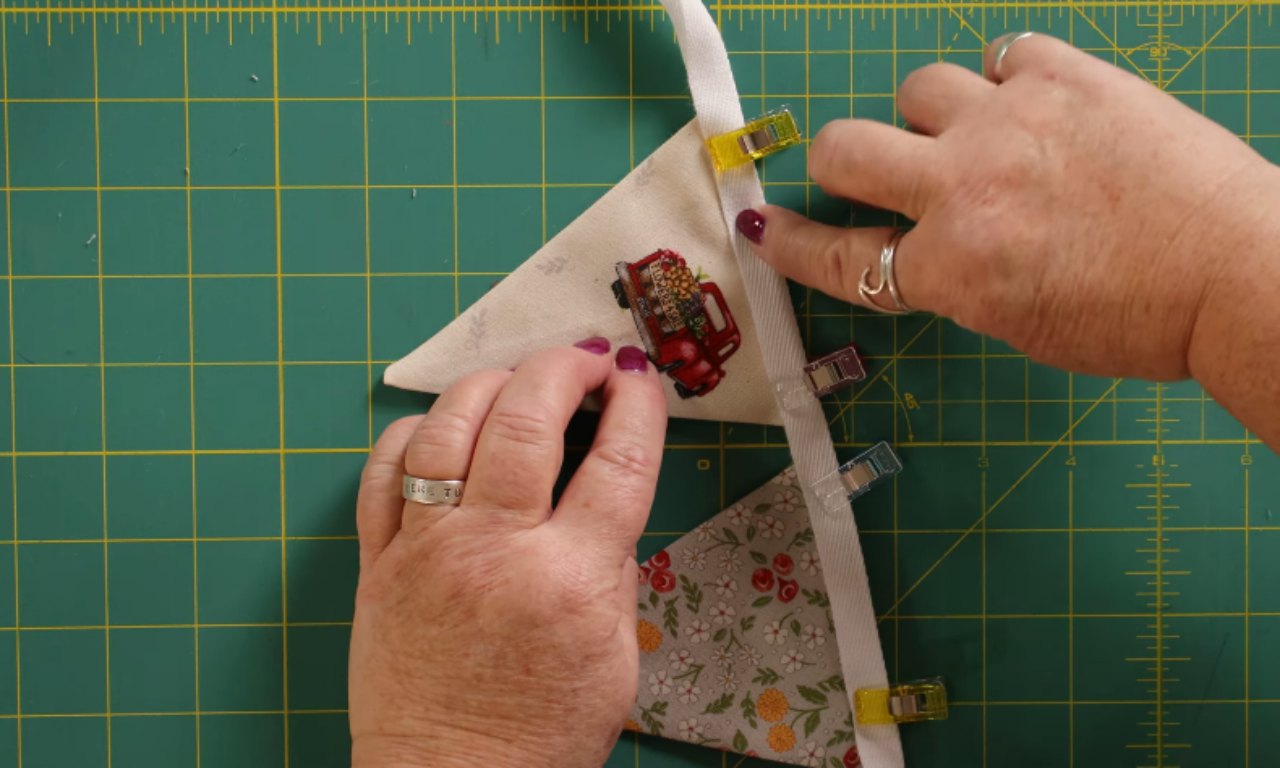 A close-up view of two triangular fabric pieces clipped to white bias tape with small colorful sewing clips. One triangle features a red vintage truck graphic, while the other has a floral pattern. A pair of hands is shown positioning the fabric with a green cutting mat underneath.
