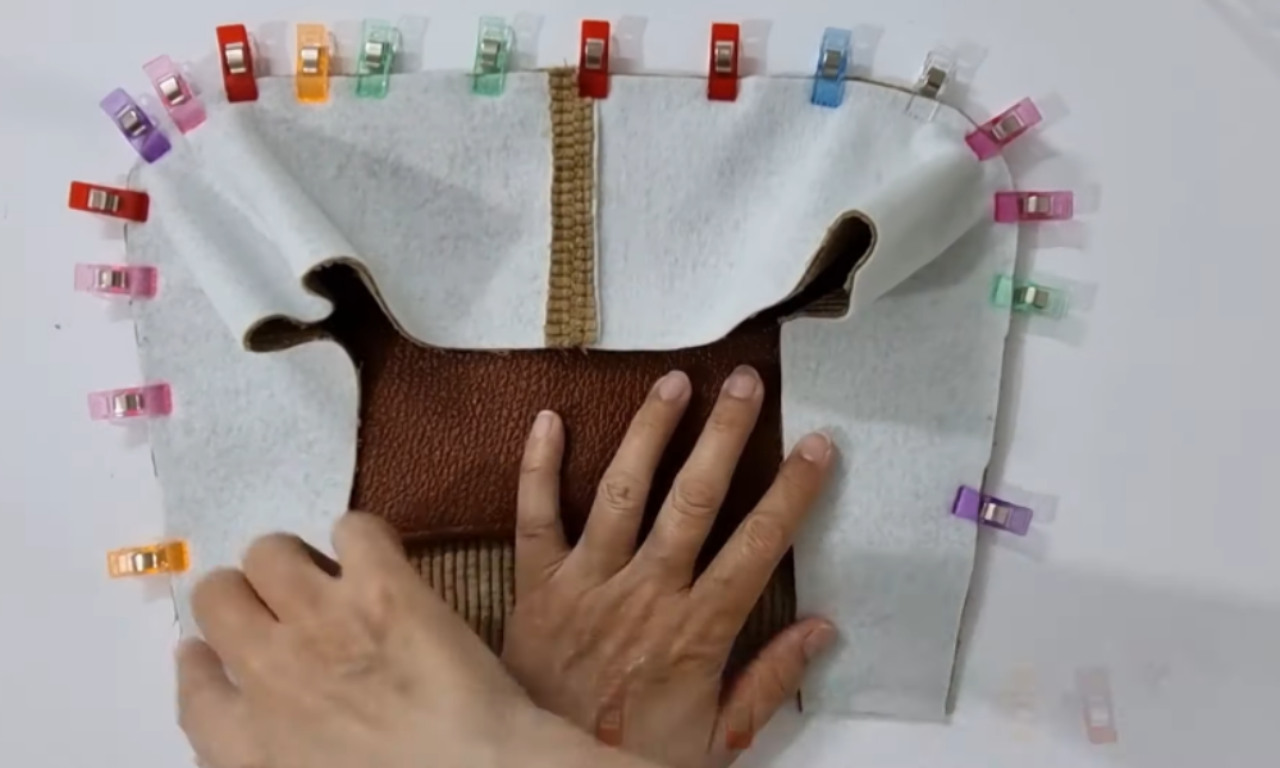 The inside of a bag is being assembled. A person's hands are pressing down brown leather and corduroy fabric layers, with white interfacing material on top. Colorful sewing clips secure the fabric layers along the edge in preparation for stitching.