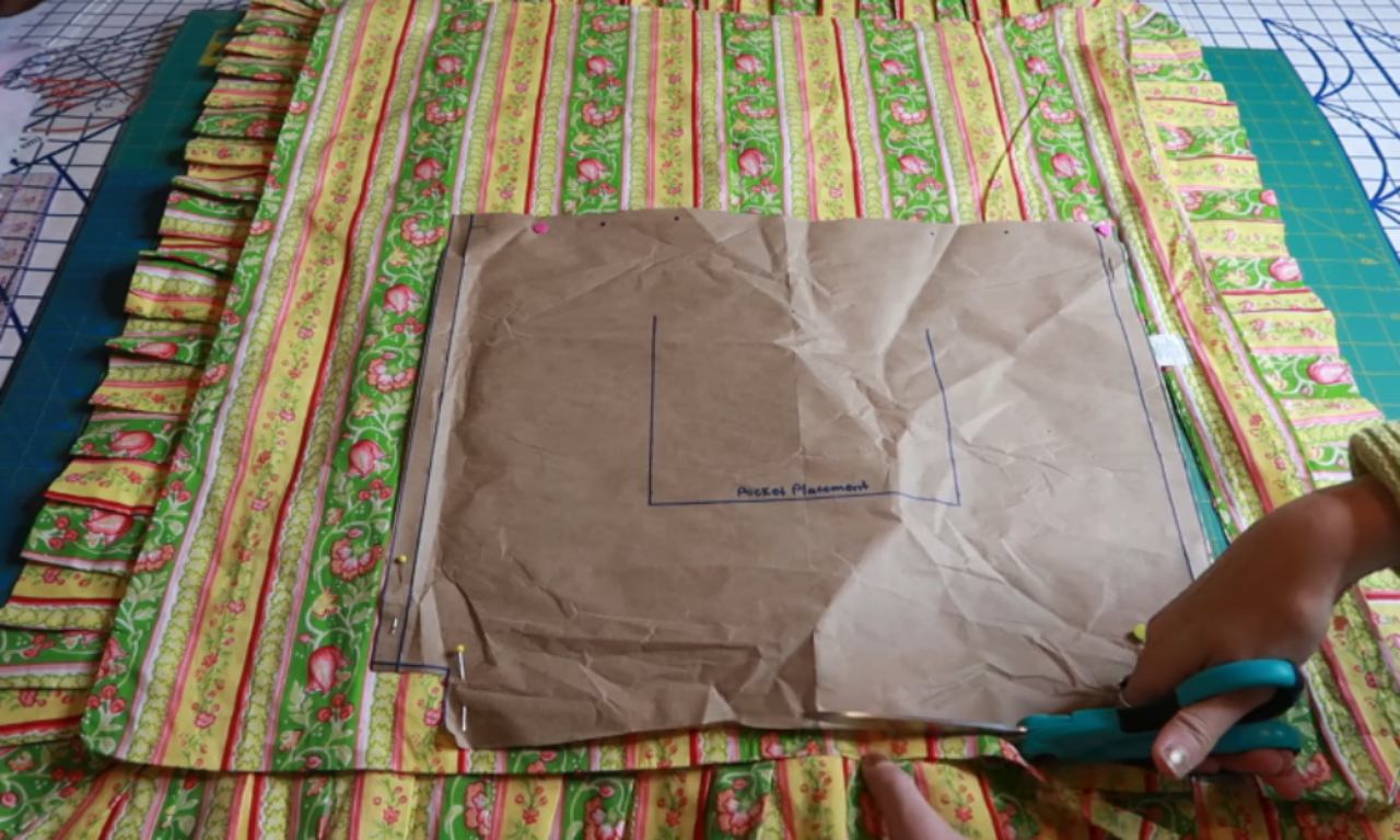 A person is cutting quilted fabric along a pinned brown paper pattern labeled "Pocket Placement." The fabric has a striped floral design in green, yellow, pink, and red tones.