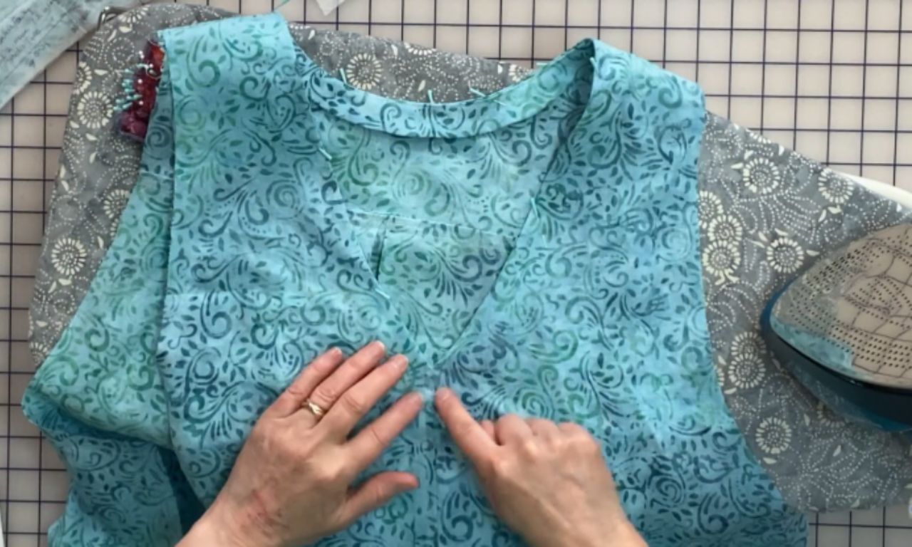 Hands pointing to a completed neckline on a blue patterned tunic with a V-neck design. The tunic is laid out flat on a pressing board with a steam iron visible to the right.