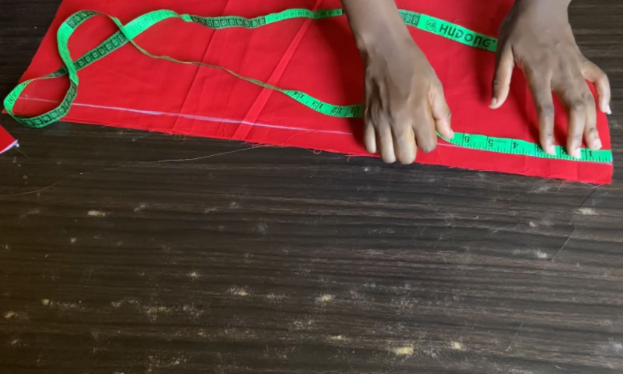 Hands measuring a folded piece of red fabric with a green measuring tape on a dark wooden table. The fabric has been marked with white lines, showing preparation for attaching the sleeve pieces to the cuff.
