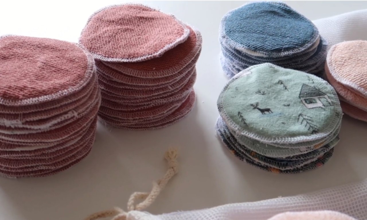 Close-up of several stacks of round, reusable fabric pads in various colors including pink, blue, green with a printed design, and peach. The pads are neatly sewn with serged edges and arranged on a white surface next to a mesh laundry bag.