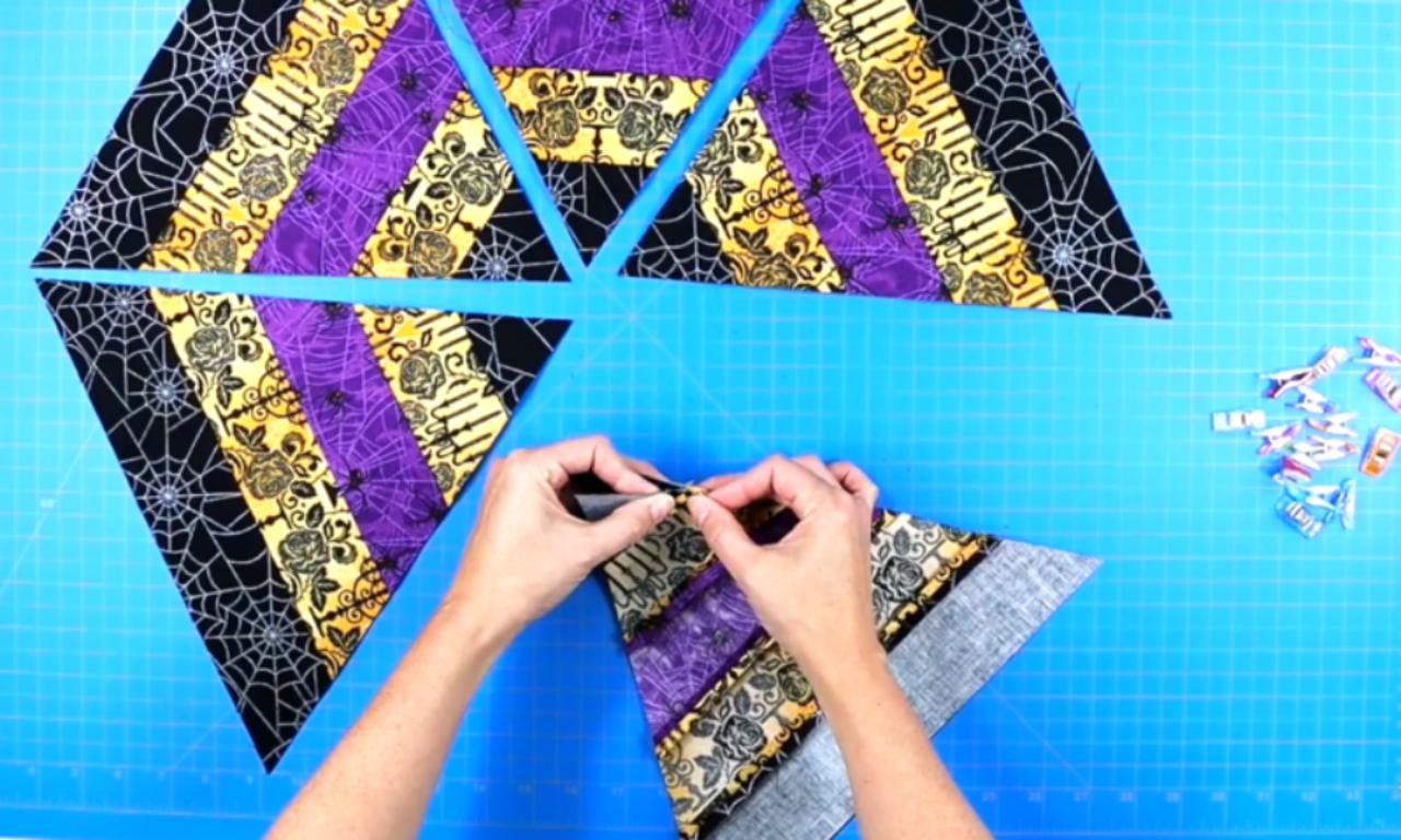 Hands are shown pinching two triangular fabric pieces together to align them before sewing. Several matching triangular pieces are arranged around the workspace, forming a partial hexagon pattern. Quilting clips are scattered on the right side over a blue cutting mat.