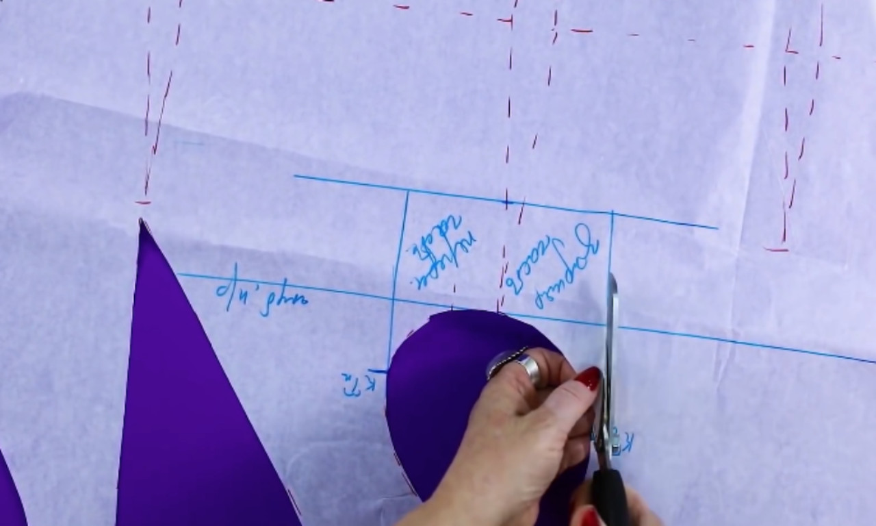 A person is cutting a pattern piece with scissors along a blue marked line on tracing paper. The paper includes handwritten labels in Russian, and part of the pattern for a sleeve or armhole is visible in a curved shape.