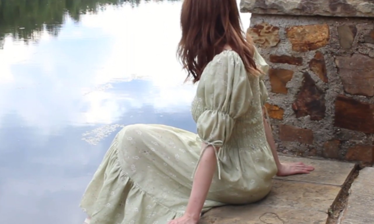 A woman in a light green, puff-sleeved dress with a smocked bodice sits on a stone ledge by a calm lake, facing away from the camera. The reflection of the sky and trees is visible in the water.