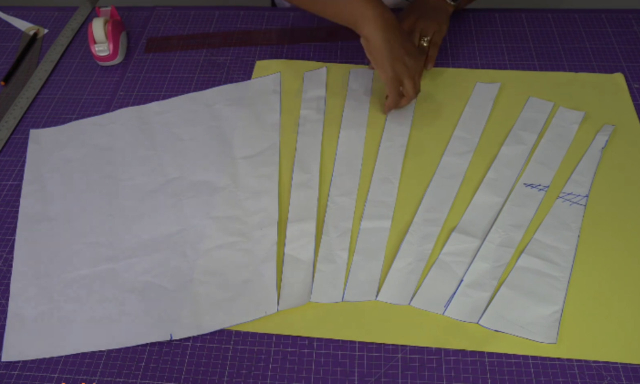 A workspace showing a person drafting a flared skirt pattern using paper. The original pencil skirt pattern is on the left, while the right side shows multiple paper segments spread apart in a fan shape on yellow paper, illustrating the slash-and-spread method to create a flounce or added volume.