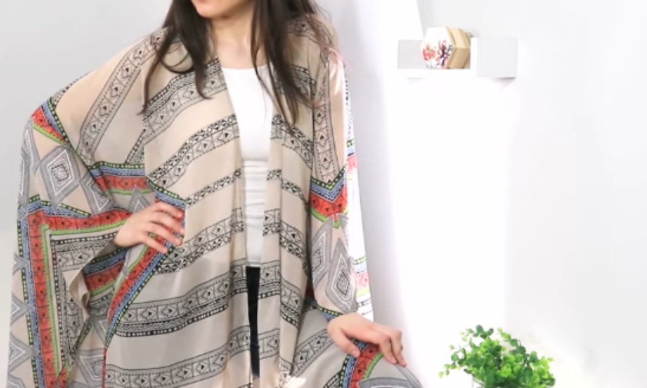 A woman is wearing a loose, patterned beach cover-up with wide sleeves. The fabric features geometric designs in black and white with accents of red, green, and blue. She poses indoors next to a white wall with a small plant and a decorative shelf in the background.