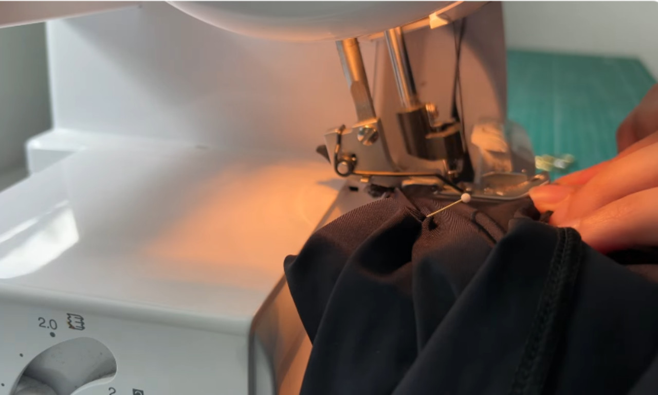 Close-up view of black fabric being sewn on a serger sewing machine. A pin holds the fabric in place while the sewing machine needle stitches through it, with warm lighting illuminating the workspace.