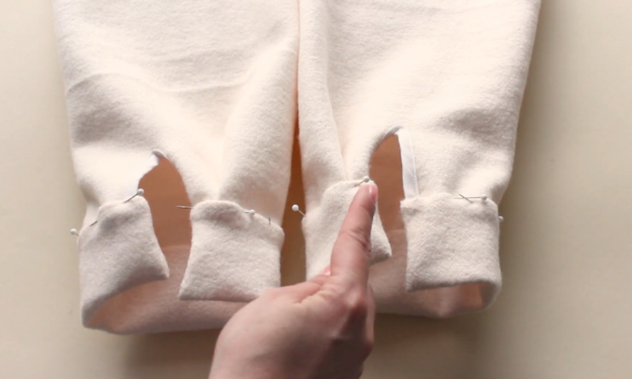 A pair of light beige sleeves pinned at the cuffs, lying flat on a surface. A person’s hand is pointing at the open seam area, illustrating the final steps of sewing and assembling the cuffs.