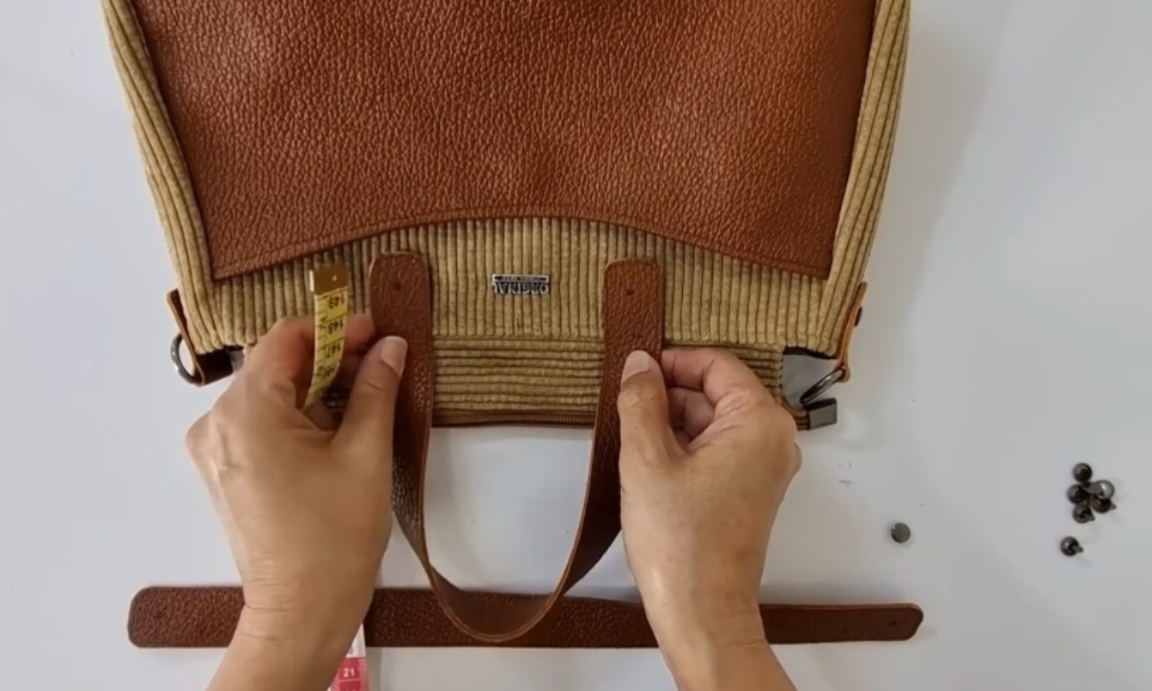 A person is measuring and positioning leather straps on a partially assembled handbag. The bag features a combination of tan corduroy fabric and brown textured leather. Small rivets and hardware are placed on the side for final assembly.
