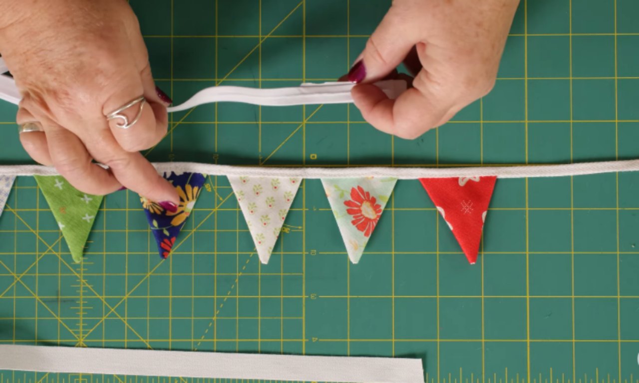 Hands are holding white bias tape above a completed bunting string with small triangle flags in various colors and floral patterns. The bunting is laid out on a green cutting mat with yellow grid lines, and the tape is being prepared for the final stitching step.