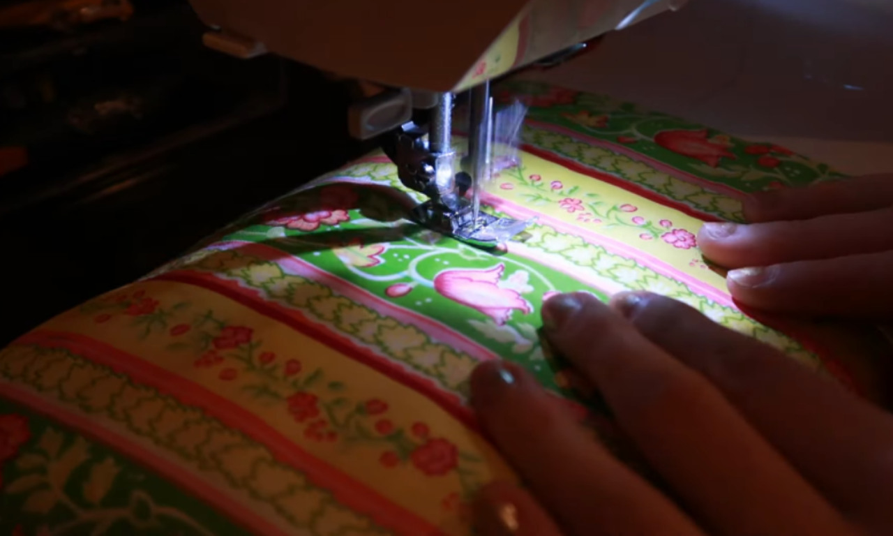 Close-up of a person using a sewing machine to stitch floral striped fabric. The hands are guiding the material under the presser foot while quilting.