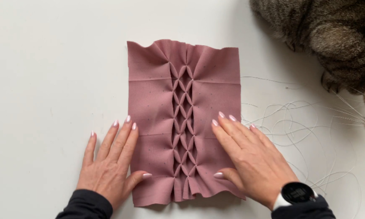Two hands spread out a partially smocked piece of dusty rose-colored fabric with a vertical strip of geometric petal-like folds in the center. A gray tabby cat is visible on the top right corner of the table, near loose threads.