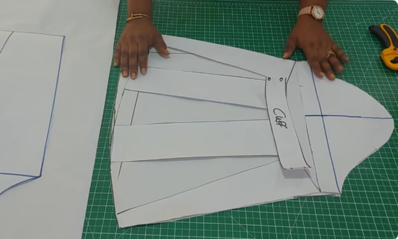 A person is holding a basic paper sleeve pattern on a green cutting mat. The pattern is slashed and spread into sections, labeled "Cuff," indicating the beginning of a bishop sleeve drafting process.