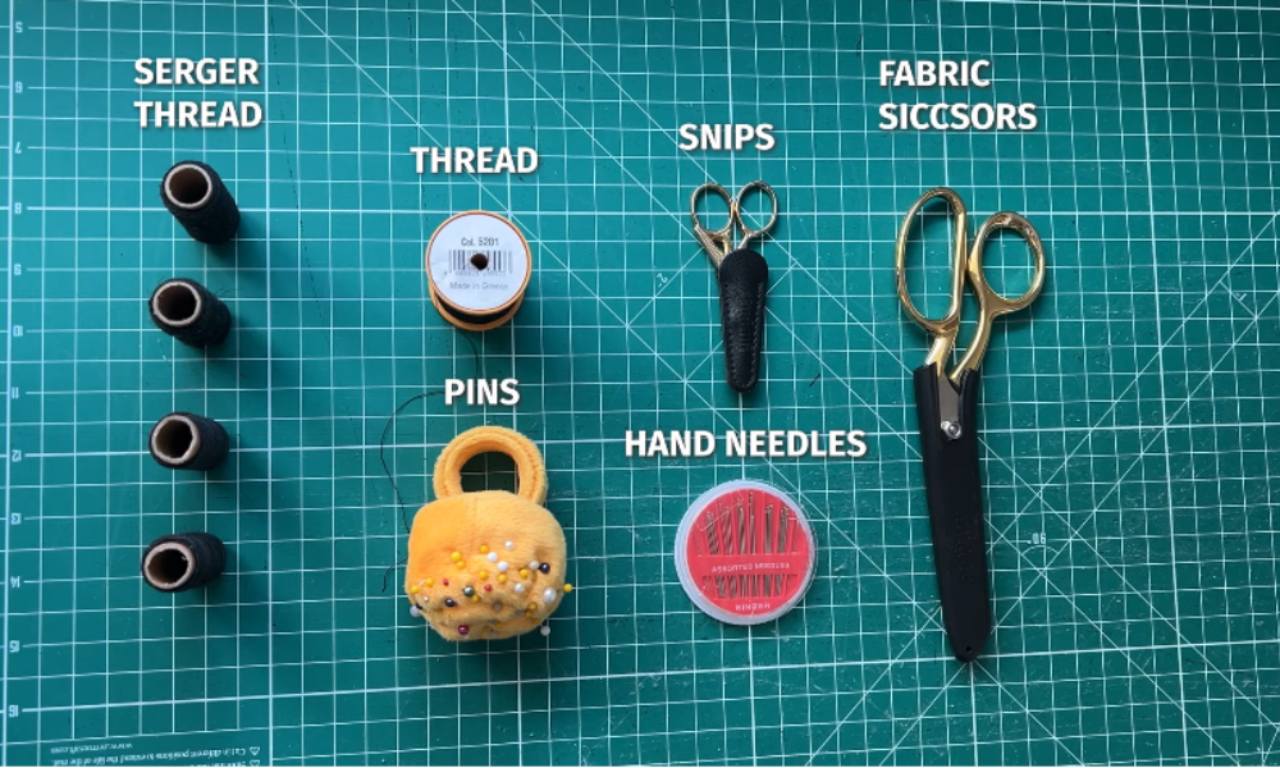 A flat lay of essential sewing tools on a green cutting mat, labeled accordingly: four cones of serger thread, a spool of regular thread, a pair of snips, fabric scissors, a pin cushion with pins, and a container of hand needles.