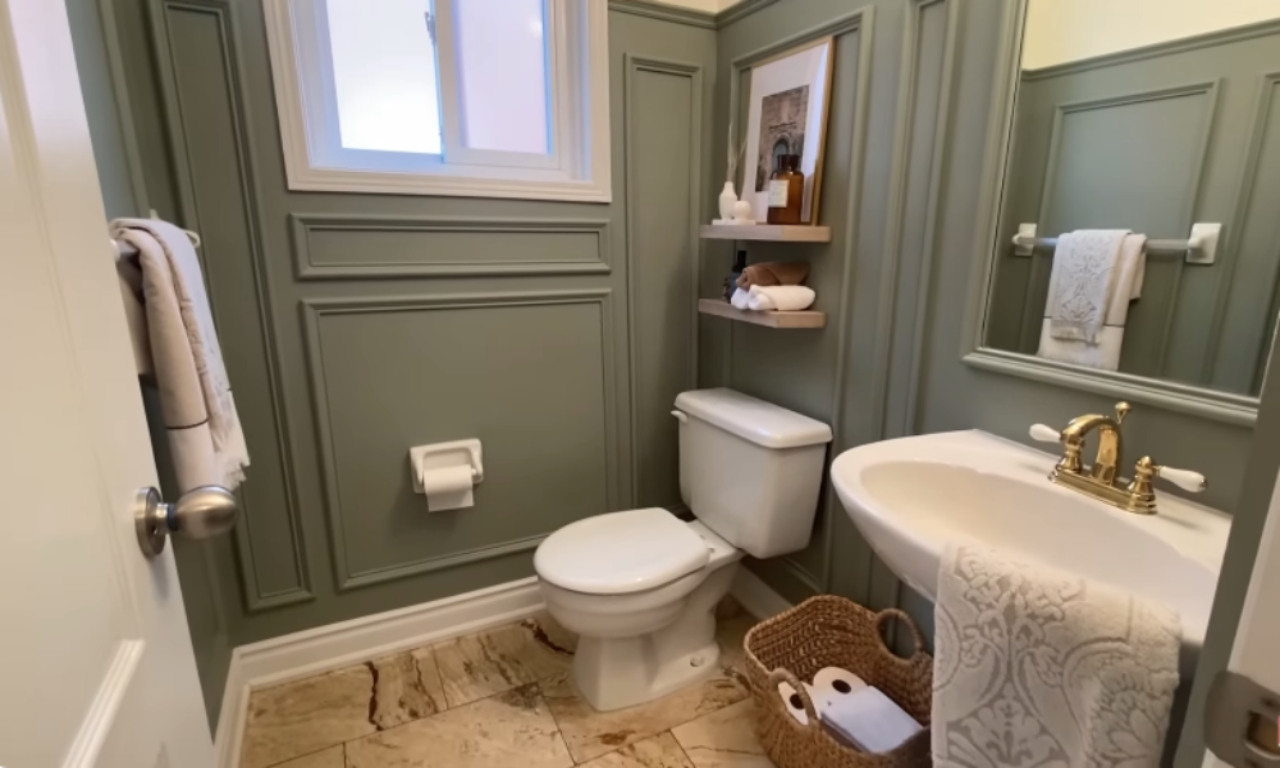 A cozy bathroom with sage green paneled walls and a neutral color palette. It includes a white toilet, a wall-mounted sink with a brass faucet, and a basket on the floor holding toilet paper and towels. Floating wooden shelves hold toiletries and framed art above the toilet, while a towel hangs on a wall-mounted rack.