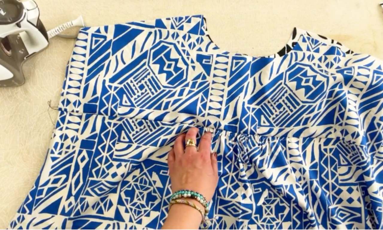 A hand holding a blue and white geometric-patterned shirt yoke that is being sewn using the burrito method. The fabric is laid flat on a beige surface with an iron placed nearby, indicating a step in garment construction.