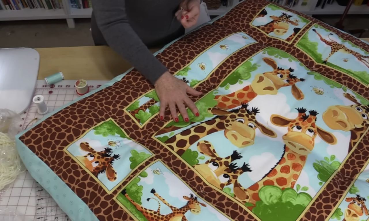 A person is smoothing out or inspecting a nearly finished quilt with bright cartoon giraffe illustrations, bordered by a giraffe-print pattern. The sewing tools from the first image are visible on the table next to the quilt.