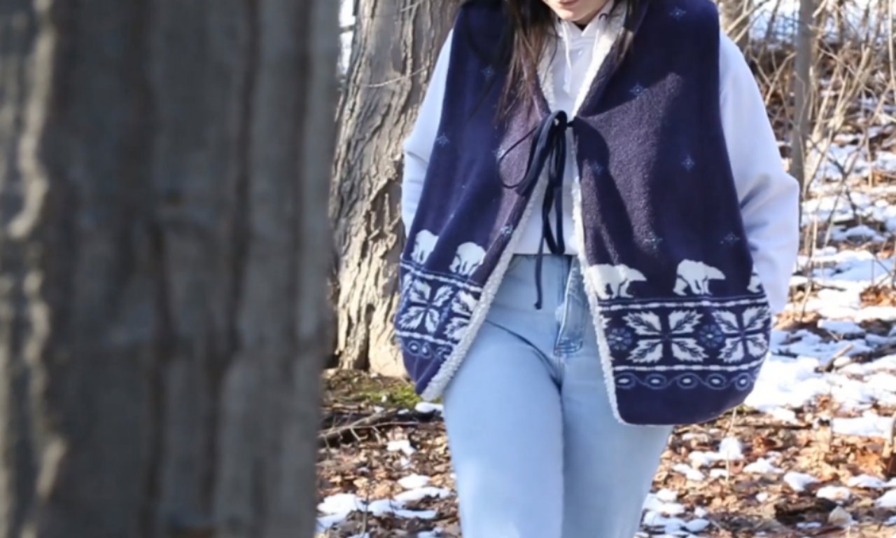 A person standing outdoors in a snowy forest wearing a handmade navy blue sherpa-lined vest with white polar bear and snowflake patterns. The vest is worn over a light hoodie and jeans.