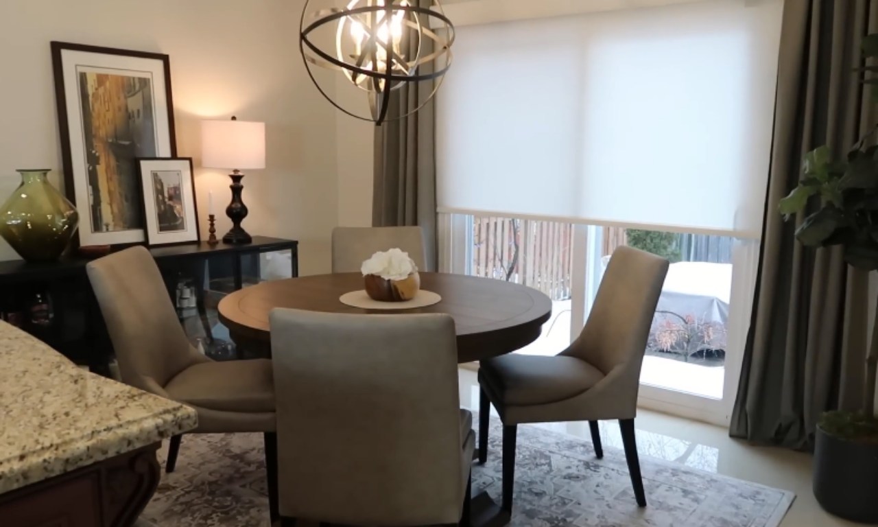Modern dining room with a round wooden table, four beige wipeable chairs, a decorative centerpiece, and a geometric chandelier above. A sideboard with framed art, lamp, and vase is against the wall, with large windows covered by white roller shades in the background.