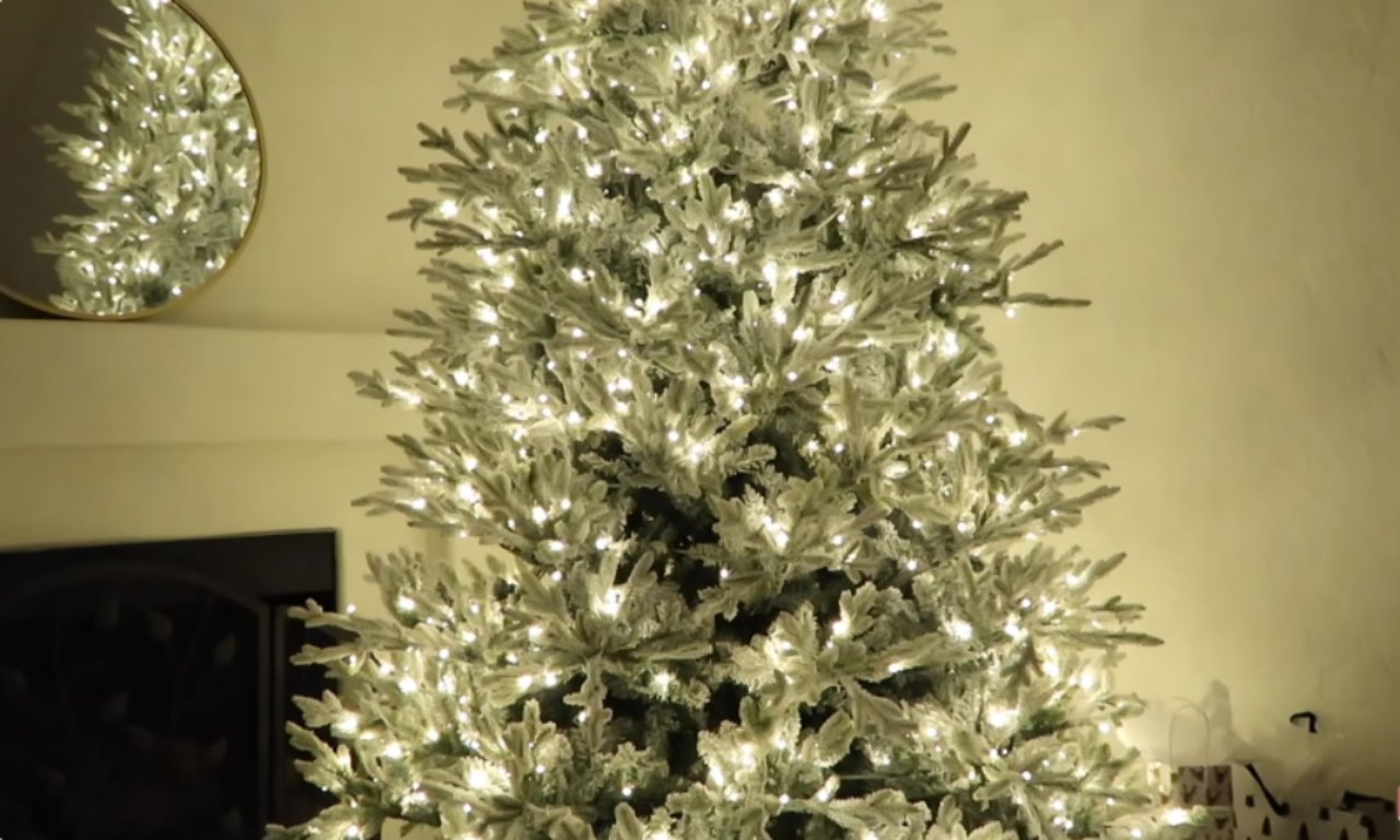 An artificial Christmas tree is decorated with a dusting of flocking and warm white lights. In the background, a circular gold mirror hangs on the wall.