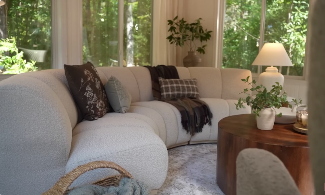 A bright and inviting living room corner with a plush curved sectional sofa in light fabric, adorned with assorted throw pillows and a plaid blanket. A round wooden coffee table holds decorative items including a candle and a vase of greenery. Large windows offer a view of lush greenery outside, and a white table lamp sits on a side table.
