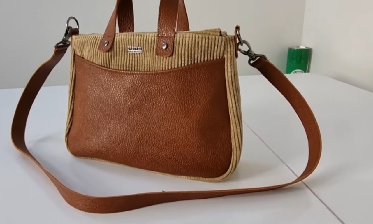 A finished handbag is displayed on a white surface. The bag is made of brown leather and tan corduroy fabric, featuring two short handles and a detachable long shoulder strap. A metallic brand label is affixed near the top center.
